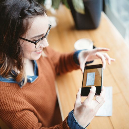 A smiling young woman takes a picture with her smart phone of a check or paycheck for digital electronic depositing, also known as "Remote Deposit Capture". She sits in a coffee shop, enjoying an espresso latte. Bright sunlight shines in the window. Horizontal image.